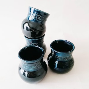 Puede incluir: Cuatro jarrones de cerámica azul oscuro con acabado brillante dispuestos sobre una superficie blanca. Cada jarrón tiene un cuerpo redondeado, una abertura ancha y una banda decorativa de líneas grabadas cerca de la parte superior. Los jarrones están apilados.