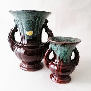 May include: Two ceramic vases with a dark brown base and a teal and green speckled top. Each vase has two ornate handles. One vase is larger than the other. A small yellow sticker is on the larger vase.