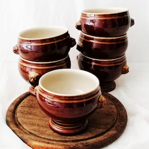 Puede incluir: Un juego de cinco cuencos de sopa de cerámica marrón con acabado brillante, interior color crema, base de pedestal y dos pequeñas asas con forma de cabeza de león en los laterales. Los cuencos están apilados y uno se sitúa en primer plano.