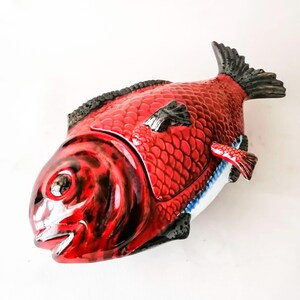 Könnte beinhalten: Ein roter Keramikbehälter in Fischform mit einem Klappdeckel. Der Fisch hat schwarze Details an den Flossen und Kanten. Der Behälter hat ein Schuppenmuster und einen weißen und blauen Bauch.