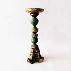 May include: An antique candlestick holder, painted green and gold. The holder features a wide, circular top, a turned stem, and a detailed base. The distressed paintwork gives it a rustic, aged appearance, suitable for a vintage aesthetic.