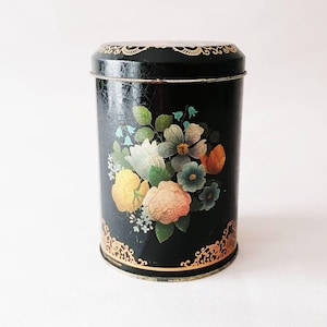 Puede incluir: Una lata cilíndrica negra con un diseño floral. La lata presenta un ramo de flores coloridas en tonos amarillos, rosas, azules y naranjas. Los detalles de filigrana dorada acentúan los bordes superior e inferior, añadiendo un toque vintage.