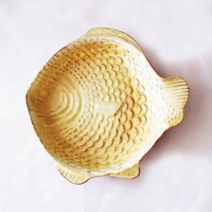 Puede incluir: Un plato de cerámica con forma de pez y un esmalte de color amarillo claro y crema. La superficie presenta un patrón de escamas texturizado y en relieve en todo el cuerpo, con líneas definidas para la cabeza, las aletas y la cola, formando un cuenco poco profundo.
