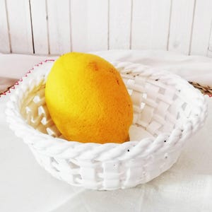 May include: A bright yellow lemon resting inside a white ceramic basket. The basket features a woven lattice design with a thick, braided rim. The surface of the lemon is textured and vibrant, contrasting with the smooth, solid white finish of the bowl.