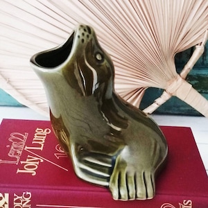 Puede incluir: Una figura de foca de cerámica en color verde oliva brillante con la boca abierta, diseñada como un recipiente decorativo. La foca está esculpida en posición sentada con detalles visibles en las aletas y un acabado liso y reflectante.
