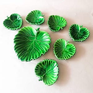Op de afbeelding: Een verzameling van acht glanzend groene keramische bladvormige schalen. De schalen variëren in grootte, met een groot centraal blad omringd door kleinere. De bladeren hebben gedetailleerde nerven en een levendige, naturalistische uitstraling.