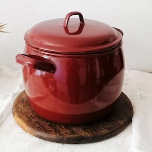 May include: A deep red enamel stockpot with a matching lid and two side handles. The pot has a smooth, glossy finish and a rounded, cylindrical body shape. The lid features a small, arched handle at the centre for easy lifting and removal.