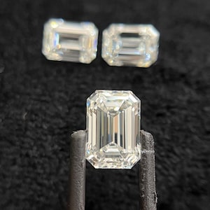 May include: Three clear, emerald-cut gemstones with a brilliant sparkle. One is held by a pair of tweezers, while two others rest above. The gems are set against a dark background.