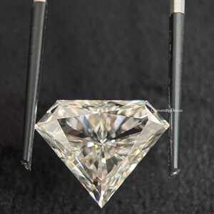 以下が含まれることがあります： 暗い背景に、金属製のピンセットで保持された、大きく透明でファセットカットされたダイヤモンド。ダイヤモンドの多くのファセットが光を反射し、鮮やかな輝きを生み出しています。「DiamondbyDenzo」の文字が右上に見えます。