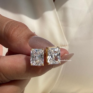 Op de afbeelding: Een paar goudkleurige vierkante oorstekers met heldere, rechthoekig geslepen stenen. De oorbellen worden vastgehouden door een hand met verzorgde nagels. De oorbellen zijn gezet in een goudkleurig metaal.