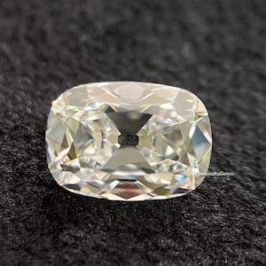 May include: A cushion-cut diamond with a brilliant, multifaceted surface. The gemstone is clear and colorless, reflecting light to create a sparkling effect. The diamond is set against a dark, textured background. The text "DiamondbyDenzo" is visible.