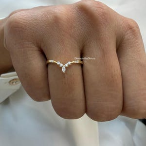 May include: A gold-colored ring with a V-shaped design. The ring features small, clear gemstones. The text "DiamondbyDenzo" is visible on the ring. The ring is on a hand.