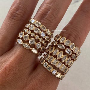 May include: A collection of gold rings featuring various diamond cuts including emerald, marquise, and round stones set in bezel settings. The rings are stacked on fingers, showcasing different geometric patterns and metal bands with the text Diamondbybenzo.