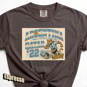 May include: Espresso-colored t-shirt featuring a graphic celebrating Argentina's World Football Championship in Qatar '22. The design includes a stylized illustration of a jaguar kicking a soccer ball, with text that reads "Argentina World Football Champions".