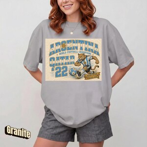 May include: A gray t-shirt featuring a graphic of a soccer-playing jaguar in an Argentina jersey, with the text "ARGENTINA QATAR '22 WORLD FOOTBALL CHAMPIONS". The shirt is a relaxed fit.