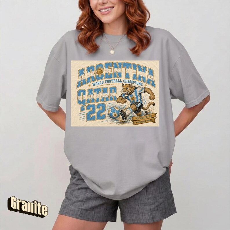 May include: A gray t-shirt featuring a graphic of a soccer-playing jaguar in an Argentina jersey, with the text "ARGENTINA QATAR '22 WORLD FOOTBALL CHAMPIONS". The shirt is a relaxed fit.