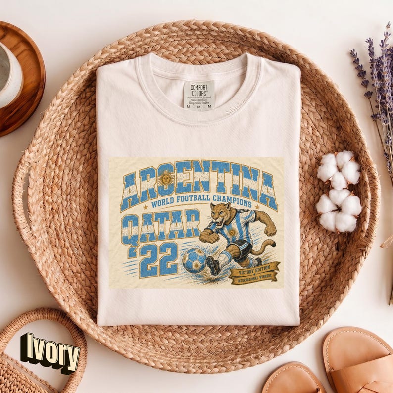 May include: Cream-colored t-shirt featuring a graphic of a cougar in an Argentina soccer uniform kicking a soccer ball. The text reads "ARGENTINA WORLD FOOTBALL CHAMPIONS QATAR '22". The shirt is displayed in a woven basket.