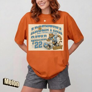 May include: Orange t-shirt with a graphic featuring the text "ARGENTINA QATAR '22 WORLD FOOTBALL CHAMPIONS" and an illustration of a cougar kicking a soccer ball. The shirt is a solid orange color.