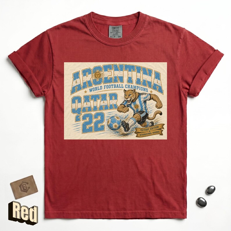 May include: Red t-shirt featuring a graphic of a puma kicking a soccer ball, with the text "ARGENTINA WORLD FOOTBALL CHAMPIONS QATAR '22". The design is in blue and white, with a vintage style.