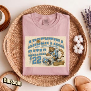 May include: A light pink t-shirt with a graphic featuring the text "ARGENTINA QATAR 22 WORLD FOOTBALL CHAMPIONS". The graphic includes a cougar in a soccer uniform kicking a soccer ball. The shirt is in a woven basket.