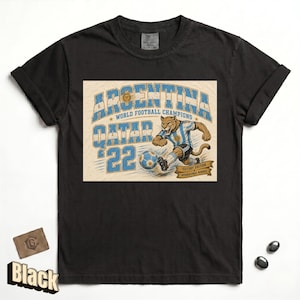 May include: Black t-shirt featuring a graphic of a puma in an Argentina soccer uniform kicking a soccer ball. The text reads "ARGENTINA WORLD FOOTBALL CHAMPIONS QATAR '22 VICTORY EDITION INTERNATIONAL WINNERS".