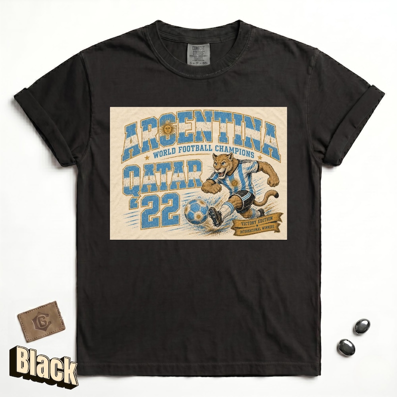 May include: Black t-shirt featuring a graphic of a puma in an Argentina soccer uniform kicking a soccer ball. The text reads "ARGENTINA WORLD FOOTBALL CHAMPIONS QATAR '22 VICTORY EDITION INTERNATIONAL WINNERS".