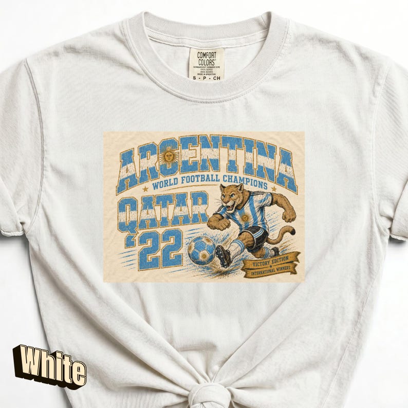 May include: White t-shirt featuring a graphic of a soccer-playing jaguar in an Argentina jersey. The design includes the text "ARGENTINA WORLD FOOTBALL CHAMPIONS QATAR '22" in blue and gold. The shirt has a crew neck and short sleeves.