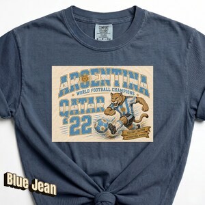 May include: Blue jean t-shirt featuring a graphic of a soccer-playing jaguar in an Argentina jersey. The graphic includes the text "ARGENTINA WORLD FOOTBALL CHAMPIONS QATAR '22". The shirt has a crew neck and short sleeves.