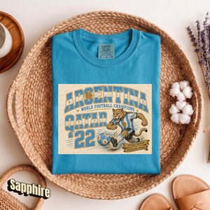 May include: Sapphire blue t-shirt with a graphic featuring the text "ARGENTINA WORLD FOOTBALL CHAMPIONS QATAR '22". The graphic includes a cougar in a soccer uniform kicking a soccer ball. The shirt is displayed in a woven basket.