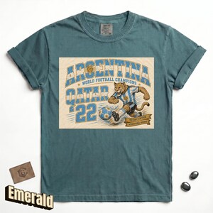 May include: Emerald green t-shirt with a graphic featuring a cougar kicking a soccer ball, the words "ARGENTINA QATAR '22 WORLD FOOTBALL CHAMPIONS" in blue and white. The shirt has rolled-up sleeves.
