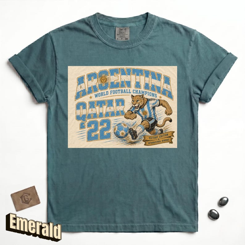 May include: Emerald green t-shirt with a graphic featuring a cougar kicking a soccer ball, the words "ARGENTINA QATAR '22 WORLD FOOTBALL CHAMPIONS" in blue and white. The shirt has rolled-up sleeves.