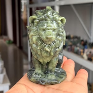 Op de afbeelding: Een gedetailleerde groene stenen leeuwensculptuur. De leeuw is gebeeldhouwd met een vloeiende manen en een koninklijke pose. De sculptuur wordt in een hand gehouden, wat de grootte en het vakmanschap benadrukt. Het thema is decoratieve kunst.