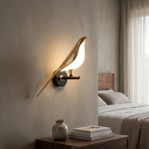 Può includere: Lampada da parete a forma di uccello con finitura dorata. La lampada ha un centro bianco brillante e una base nera. L'uccello è posato su un'asta nera, creando un'illuminazione moderna ed elegante.
