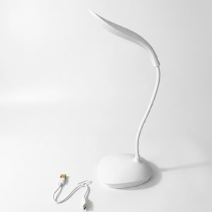 May include: A white LED desk lamp with a leaf-shaped light head and a curved, flexible neck. The lamp has a round base and includes a USB charging cable. The design is minimalist and modern, suitable for a desk or bedside table.