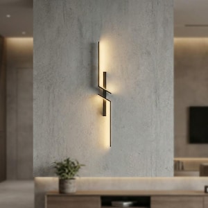 Può includere: Applique da parete nero moderno con design geometrico. Ha una forma rettangolare verticale con linee intersecanti ed emette una luce calda. Montato su una parete grigia strutturata, aggiunge un tocco contemporaneo allo spazio.