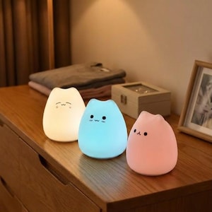 May include: Three cat-shaped night lights in white, blue, and pink. Each light has a simple face design. The lights are on a wooden surface, with a wooden box and framed picture in the background.