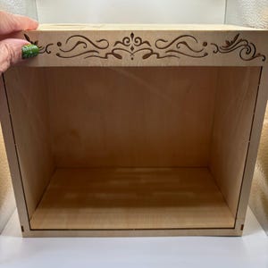 May include: A light-coloured wooden box with an open front. The top edge features a decorative, laser-cut design. The box has a simple, rectangular shape, with visible wood grain. The interior is empty, suggesting it's designed for display or storage.