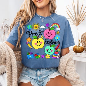 May include: A blue short-sleeve t-shirt featuring a colorful graphic print with the text Pre-2nd Explorers. The design includes three smiling apples in pink, yellow, and green, along with a globe, sun, rainbow, pencil, crayons, and books.