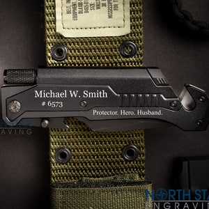 May include: Black tactical knife with a green and brown camouflage belt. The knife has a black handle and a silver blade. The knife is engraved with the text "Michael W. Smith #6573 Protector. Hero. Husband."