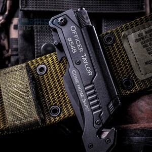 May include: Black multi-tool knife with the engraved text "OFFICER TAYLOR #548 COME HOME SAFE!" resting on a green nylon belt. The belt has a tag with text including "WAIST UNDER 30 INCHES". The background includes a boot and the North Star Engraving logo.