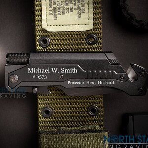 May include: Black multi-tool with the engraved name "Michael W. Smith" and the text "Protector. Hero. Husband." The tool is on a green belt with the text "Waist Under 30 inches". A brown boot and a camouflage pouch are also visible.