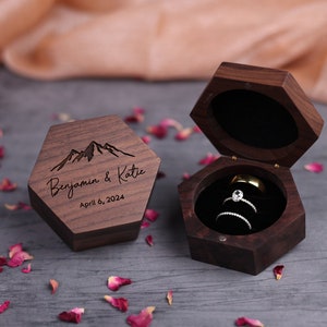 May include: A hexagonal wooden ring box with a mountain range design and the text "Benjamin & Katie April 6, 2024". The box is open and contains two rings.