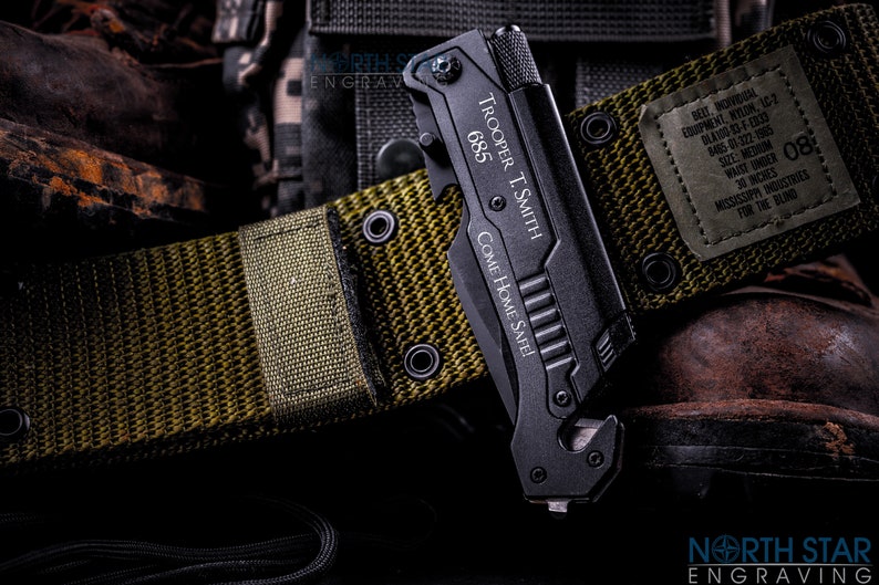 May include: Black multi-tool knife with the text "TROOPER T. SMITH 685 COME HOME SAFE!" on a green belt. The belt has a label with text including "WAIST UNDER 30 INCHES". The knife is next to a brown boot.