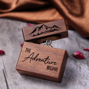 May include: A wooden ring box with a mountain scene engraved on the lid. The box is open and a ring is inside. The bottom of the box says "The Adventure Begins".