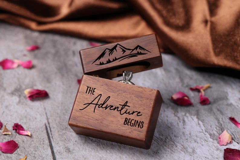May include: A wooden ring box with a mountain scene engraved on the lid. The box is open and a ring is inside. The bottom of the box says "The Adventure Begins".