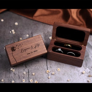 May include: A wooden ring box with a dandelion design and the text "Benjamin & Katie June 22, 2024". The box is open and two rings are visible inside.