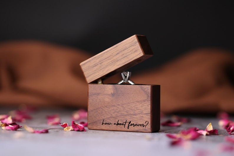 May include: A wooden ring box with the lid open, revealing a silver ring with a diamond. The box is engraved with the text "how about forever?" and is surrounded by pink rose petals.
