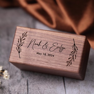 May include: A wooden ring box with a personalized inscription that reads "Noah & Evelyn May 18, 2024". The box is decorated with engraved floral designs.