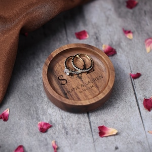May include: A wooden ring dish with the inscription "Shane & Lauren 10-18-2024" and a floral design. Two gold rings are resting on the dish.