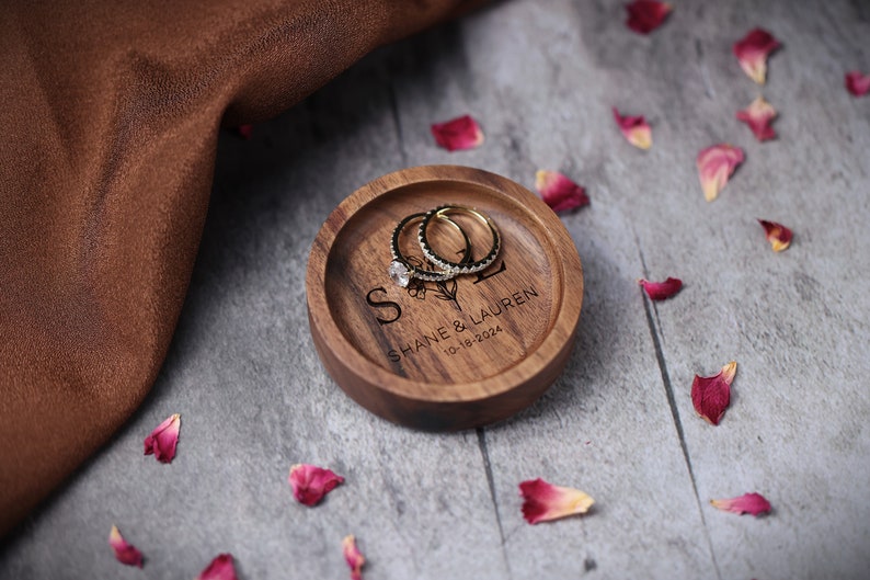 May include: A wooden ring dish with the inscription "Shane & Lauren 10-18-2024" and a floral design. Two gold rings are resting on the dish.